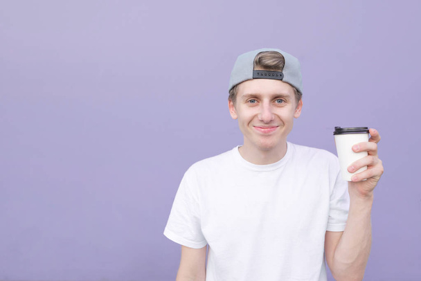 Смішний молодий чоловік одягнений у білу футболку і стоїть на пастельно-фіолетовому тлі і дивиться на камеру. Молодий чоловік зі склянкою кави на фоні фіолетової стіни
 - Фото, зображення