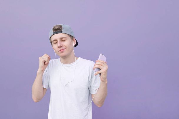 Cute young man in a white T-shirt listens to music in his headphones with his eyes closed on a pastel purple background. Pleasure of listening to music. Dance to music in the purple background - Photo, Image