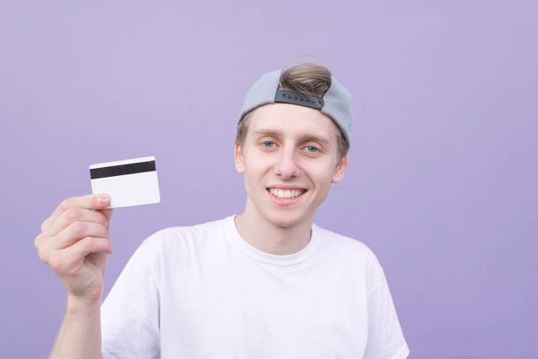 Mutlu genç adam beyaz tişört içinde elleri ve gülümser bir banka kartı mor bir arka plan üzerinde tutar. Mor bir arka plan üzerinde bir kredi kartı ile gülümseyen öğrenci - Fotoğraf, Görsel