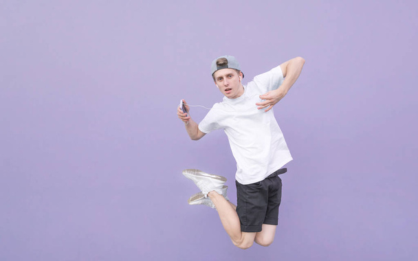 Jonge man springt met hoofdtelefoons en muziek op een pastel paars achtergrond. Portret van een tiener in vlucht met hoofdtelefoons en een smartphone op een paarse muur - Foto, afbeelding