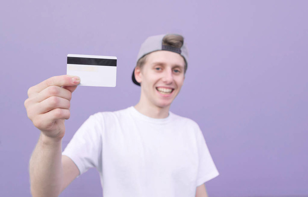 Lächelnder junger Mann hält eine Bankkarte in der Hand und lächelt auf pastelllila Hintergrund. glücklicher Student mit Kreditkarte vor dem Hintergrund einer violetten Wand. Fokus auf eine Bankkarte - Foto, Bild
