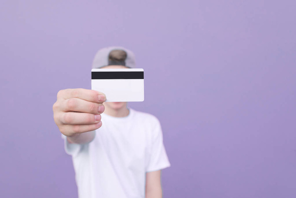Joven en una camiseta blanca con una tarjeta bancaria, su mano está sobre un fondo pastel púrpura. El hombre muestra una tarjeta de crédito en una cámara. Tarjeta bancaria en mano enfoque de primer plano
 - Foto, Imagen