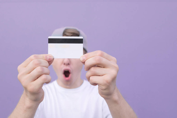 Close-up de um cartão bancário nas mãos de um homem emocional em um fundo púrpura pastel. Cartão de crédito em foco nas mãos. Espaço de cópia
 - Foto, Imagem