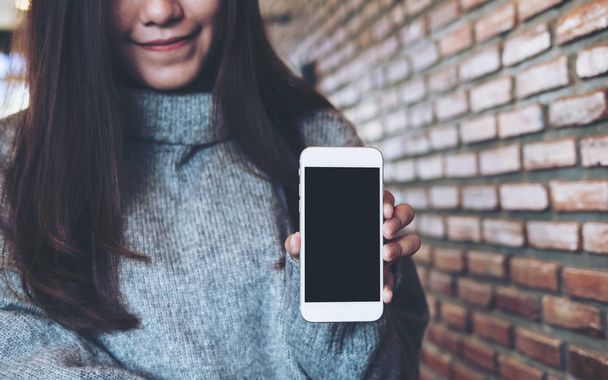 Imagen burlona de una hermosa mujer asiática sosteniendo y mostrando el teléfono móvil blanco con pantalla en blanco negro en la cafetería con fondo de pared de ladrillo
 - Foto, Imagen