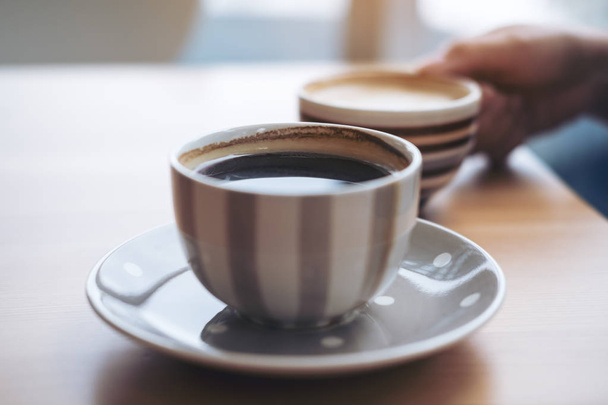 Closeup image of Americano coffee cup and woman's hand holding latte coffee cup on vintage wooden table in cafe - Photo, Image