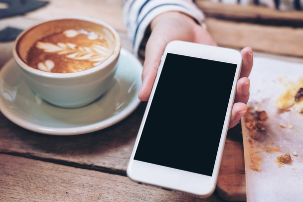 Изображение женщины, держащей и показывающей белый мобильный телефон с чистым черным экраном и чашкой кофе с латте и закусками на деревянном столе в кафе
 - Фото, изображение