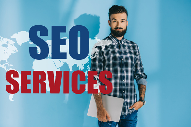 bearded developer in checkered shirt holding laptop, on blue with world map and SEO services - Photo, Image