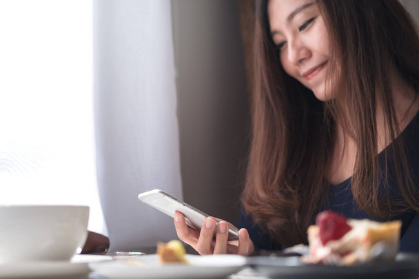 Immagine ravvicinata di una bella donna asiatica sorridente che tiene, usando e guardando smart phone con tazza di caffè bianco e piatto da dessert su tavolo in legno in caffè vintage
 - Foto, immagini