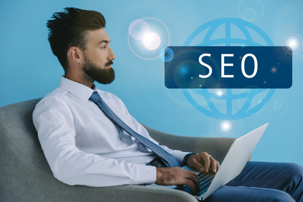 desenvolvedor barbudo usando laptop e sentado em poltrona, em azul com sinal de SEO
 - Foto, Imagem