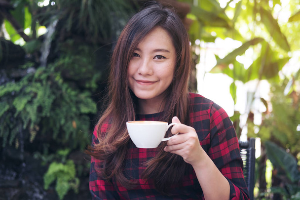 Closeup portrait image of a beautiful Asian woman holding a white mug and drinking hot coffee with feeling happy in green nature and waterfall garden background - Foto, Imagem