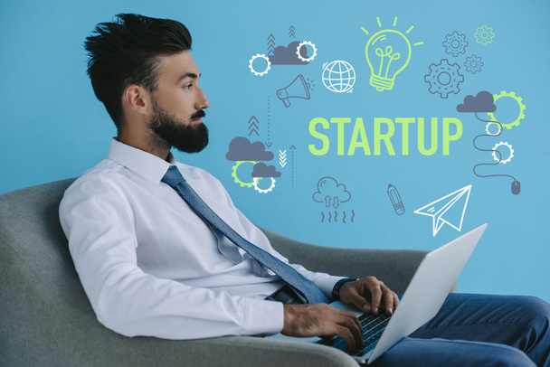 homem de negócios barbudo usando laptop e sentado em poltrona, em azul com ícones de inicialização e lâmpada
 - Foto, Imagem
