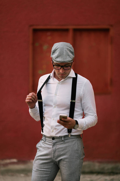 Fashionable retro dressed man wearing cap and eyeglasses on city street and using phone - 写真・画像