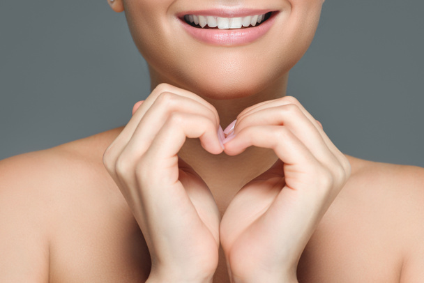 обрезанный снимок улыбающейся женщины с белыми зубами, показывающий знак сердца с руками, изолированными на сером
 - Фото, изображение