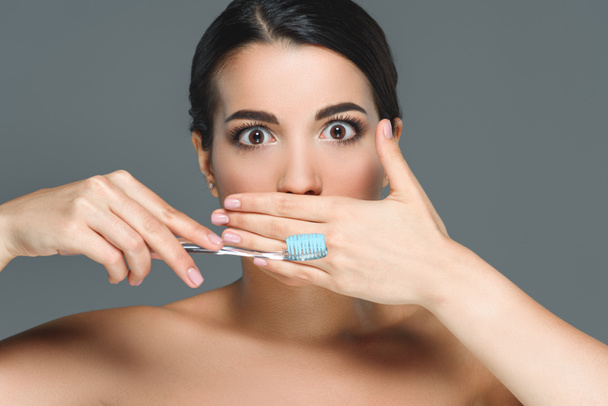 brunette woman with bare shoulders and tooth brush covering mouth isolated on grey - Fotó, kép