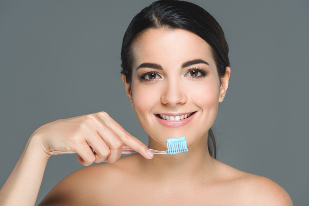 портрет улыбающейся женщины, держащей зубную щетку с зубной пастой, изолированной на сером
 - Фото, изображение