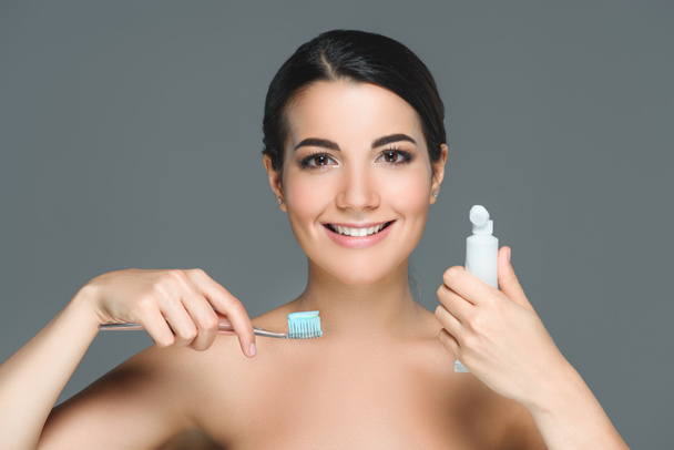 retrato de mulher sorridente com escova de dentes e tubo de pasta de dente em mãos isoladas em cinza
 - Foto, Imagem