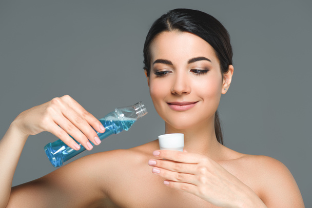portrait of smiling woman with bare shoulders holding mouthwash isolated on grey - Foto, afbeelding
