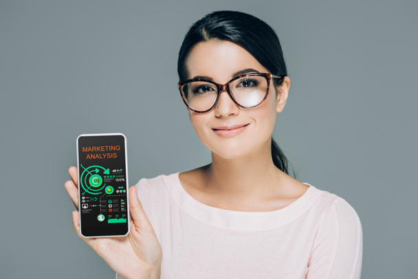 Πορτρέτο γυναίκας χαμογελαστά σε γυαλιά δείχνει smartphone με ανάλυση στην οθόνη που απομονώνονται σε γκρι μάρκετινγκ - Φωτογραφία, εικόνα