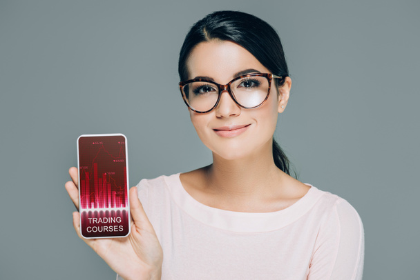 portrait of smiling woman in eyeglasses showing smartphone with trading courses logo on screen isolated on grey - Photo, Image