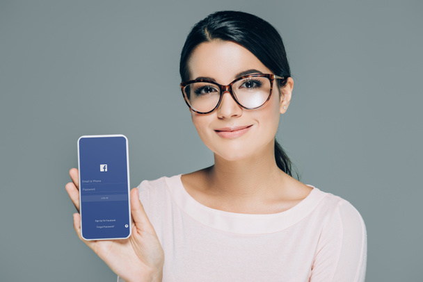 Portret kobiety uśmiechający się w okulary Wyświetlono smartphone z logo Facebooka na ekranie na szarym tle - Zdjęcie, obraz