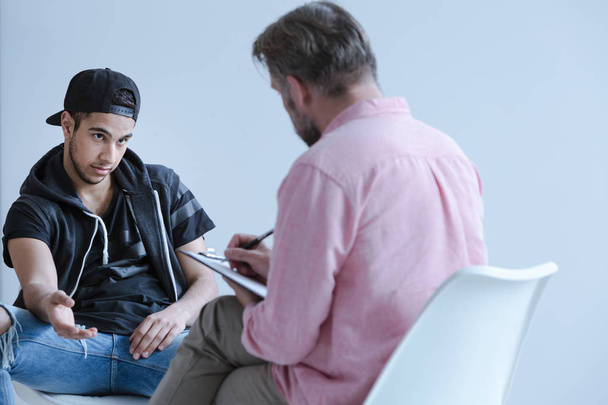 Back view of a psychology specialist talking to a rebel juvenile (front view) with violence and drugs problems during an individual counseling session. - Foto, Imagen
