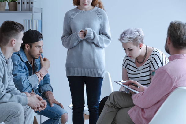 Shy withdrawn teenage girl standing and sharing her story with other participants of a group therapy during a meeting with their counselor. - Φωτογραφία, εικόνα