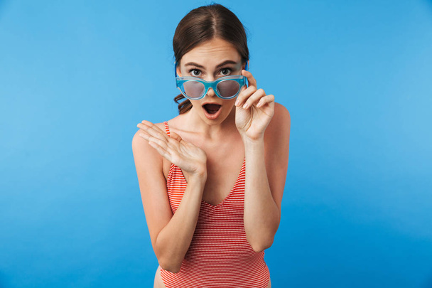 Portrait of a surprised young girl in swimsuit wearing sunglasses looking at camera isolated over blue background - Photo, Image