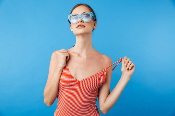 Portret van een stijlvolle jonge meisje in zwembroek dragen van een zonnebril poseren geïsoleerd op blauwe achtergrond - Foto, afbeelding