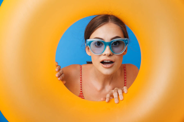 Portrait of a cheerful young girl in swimsuit wearing sunglasses looking through inflatable ring hole isolated over blue background - Fotografie, Obrázek