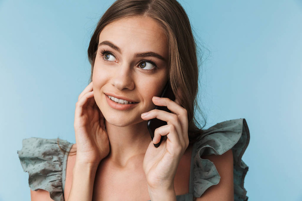 Picture of happy young woman standing isolated over blue background looking aside talking by mobile phone. - Foto, Bild
