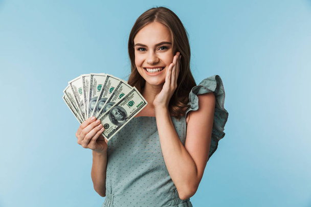 Portrait of an excited young woman in dress holding money banknotes isolated over blue background - Foto, immagini