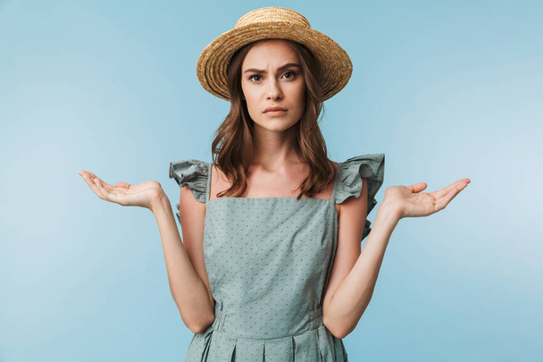 Close up portrait of a confused woman in dress and summer hat shrugging shoulders isolated over blue background - Fotoğraf, Görsel
