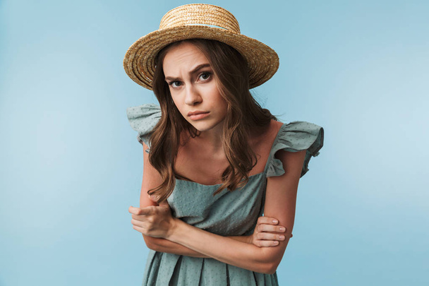 Portrait of a curious woman in dress and summer hat looking closely at camera isolated over blue background - Zdjęcie, obraz