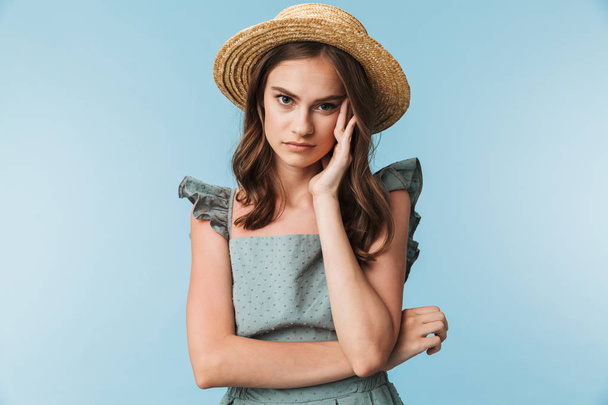 Close up portrait of a tired woman in dress and summer hat looking at camera isolated over blue background - Foto, imagen