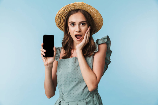 Portrait of an excited young woman in dress and summer hat showing blank screen mobile phone isolated over blue background - Foto, afbeelding