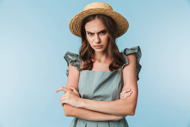 Close up portrait of an upset woman in dress and summer hat looking at camera isolated over blue background - Foto, Imagem