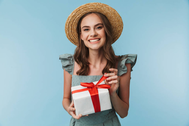 Portrait of a cheerful woman in dress and summer hat holding present box isolated over blue background - Фото, зображення