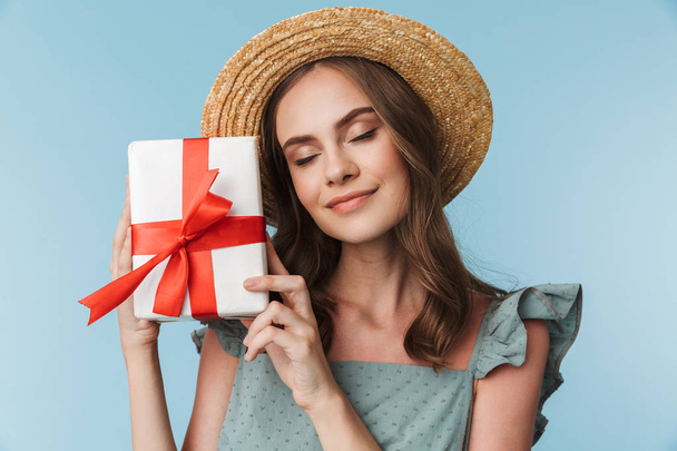 Portrait of a lovely young woman in dress and summer hat holding present box isolated over blue background - Foto, imagen