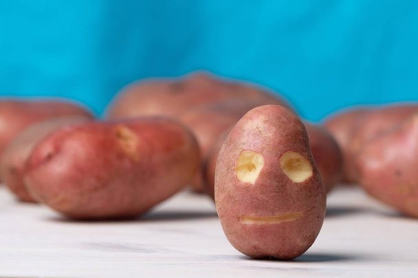 πρόσωπο σκαλισμένο σε πατάτες σε μπλε φόντο, πολλή πατάτες στο σκηνικό φόντο - Φωτογραφία, εικόνα