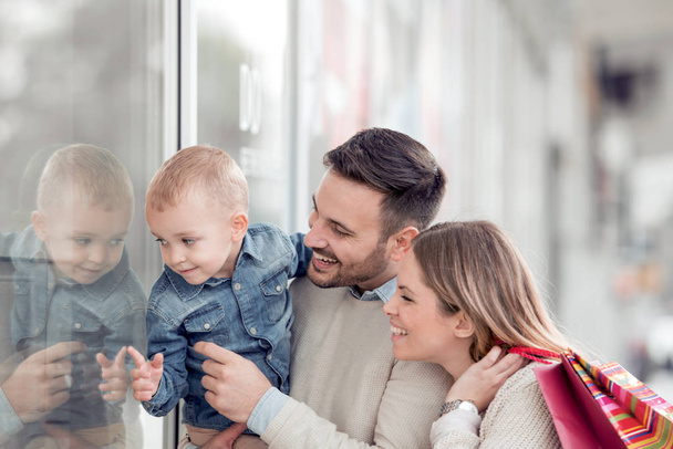 販売、消費者と家族のコンセプト-幸せな若い家族とショッピング バッグや市の店の窓を見て、彼らの息子. - 写真・画像