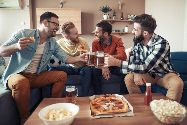 Four male friends watching TV at home while eating pizza and drinking some beer. - Photo, Image
