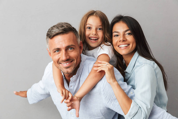 Portrait of a happy family father, mother, little daughter having fun together, father holding little daughter on his back isolated over gray background - Foto, Imagem