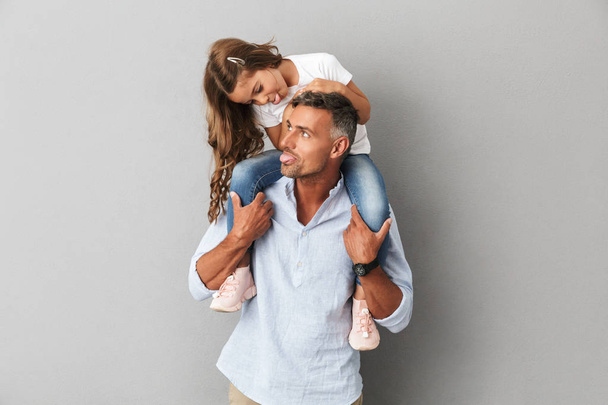 Portrait of happy family little girl and adult man having fun while daughter sitting on the neck of her father isolated over gray background - Photo, Image