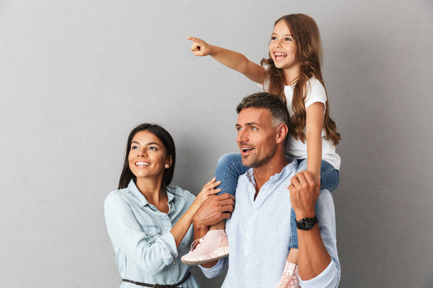 Photo of joyful family woman and man smiling and looking aside while pretty girl sitting on the neck of her happy father isolated over gray background - Photo, image