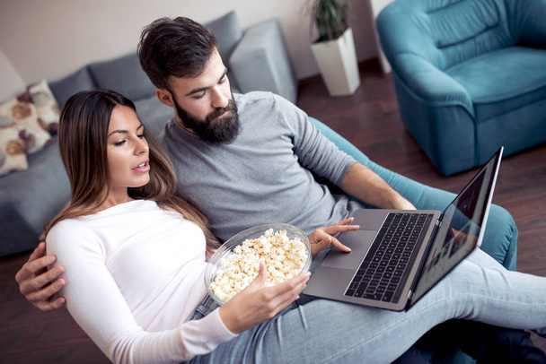 Happy young couple sitting on the sofa at home with popcorn and watching movie on laptop. - Photo, Image