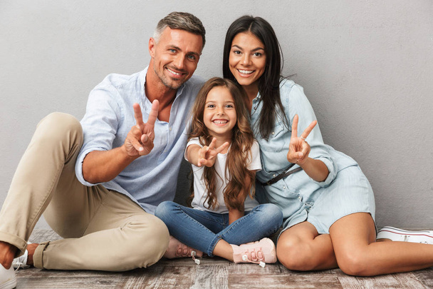 Portrait of a happy family father, mother, little daughter hugging while sitting together and showing peace gesture isolated over gray background - Fotografie, Obrázek
