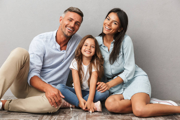Portrait of a happy family father, mother, little daughter hugging while sitting together and looking at camera isolated over gray background - Photo, Image