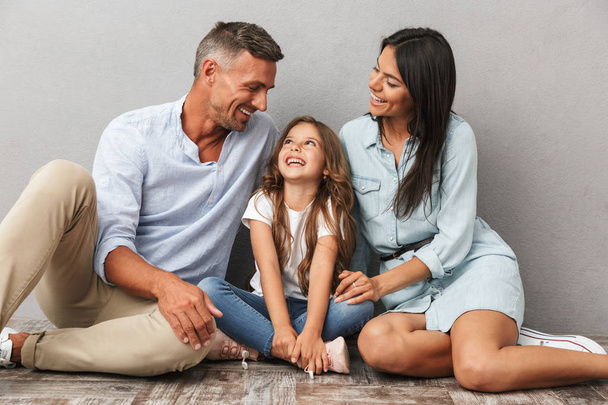 Portrait of a happy family father, mother, little daughter hugging while sitting together and looking at each other isolated over gray background - Foto, Imagem