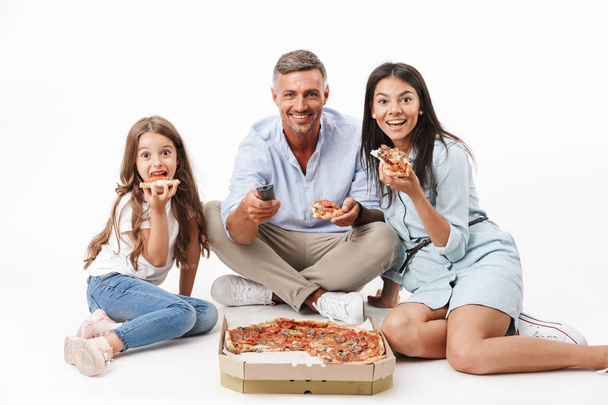 Portrait of an excited family father, mother, little daughter having fun while eating pizza and watching TV isolated over gray background - Φωτογραφία, εικόνα