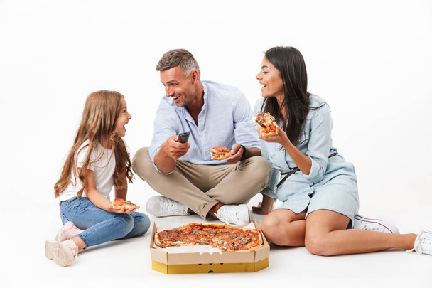 Portrait of a cheerful family father, mother, little daughter having fun while eating pizza and watching TV isolated over gray background - Valokuva, kuva